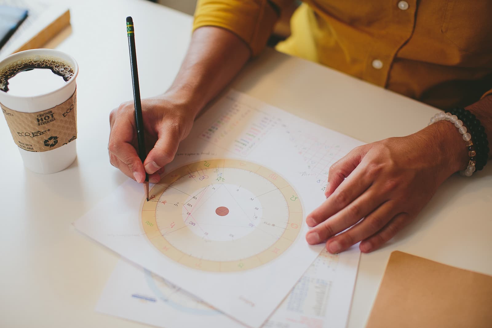 Hand annotating a printed birth chart with a pencil, coffee cup nearby
